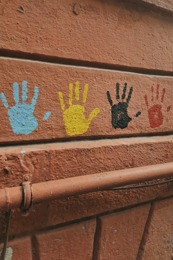 Vier bunte Handabdrücke in Blau, Gelb, Schwarz und Rot auf einer terrakottafarbenen Hauswand
